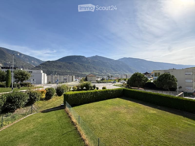 Well-maintained lawn surrounded by hedges, with a view of buildings and mountains in the background.
