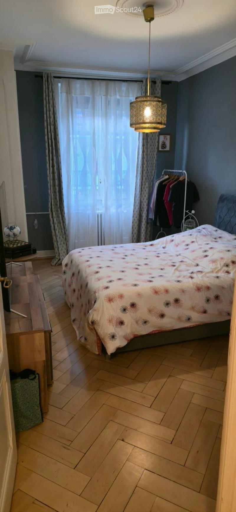 A well-lit bedroom with wooden flooring, a large window with curtains, a bed with a patterned duvet, a hanging pendant light, a wardrobe, and a nightstand with a picture frame.