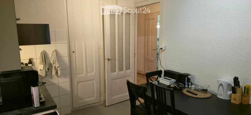 A small, modern kitchen area with a black countertop, microwave, toaster, electric kettle, and a small dining table with chairs. A screen is mounted on the wall, and a doorway leads to another room.