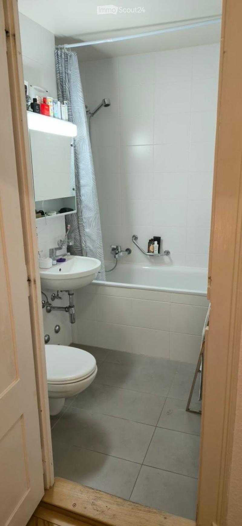 Modern bathroom with a white toilet, sink, bathtub, and shower. Features a grey tiled floor, white tiled walls, a wooden shelf, and a grey shower curtain.