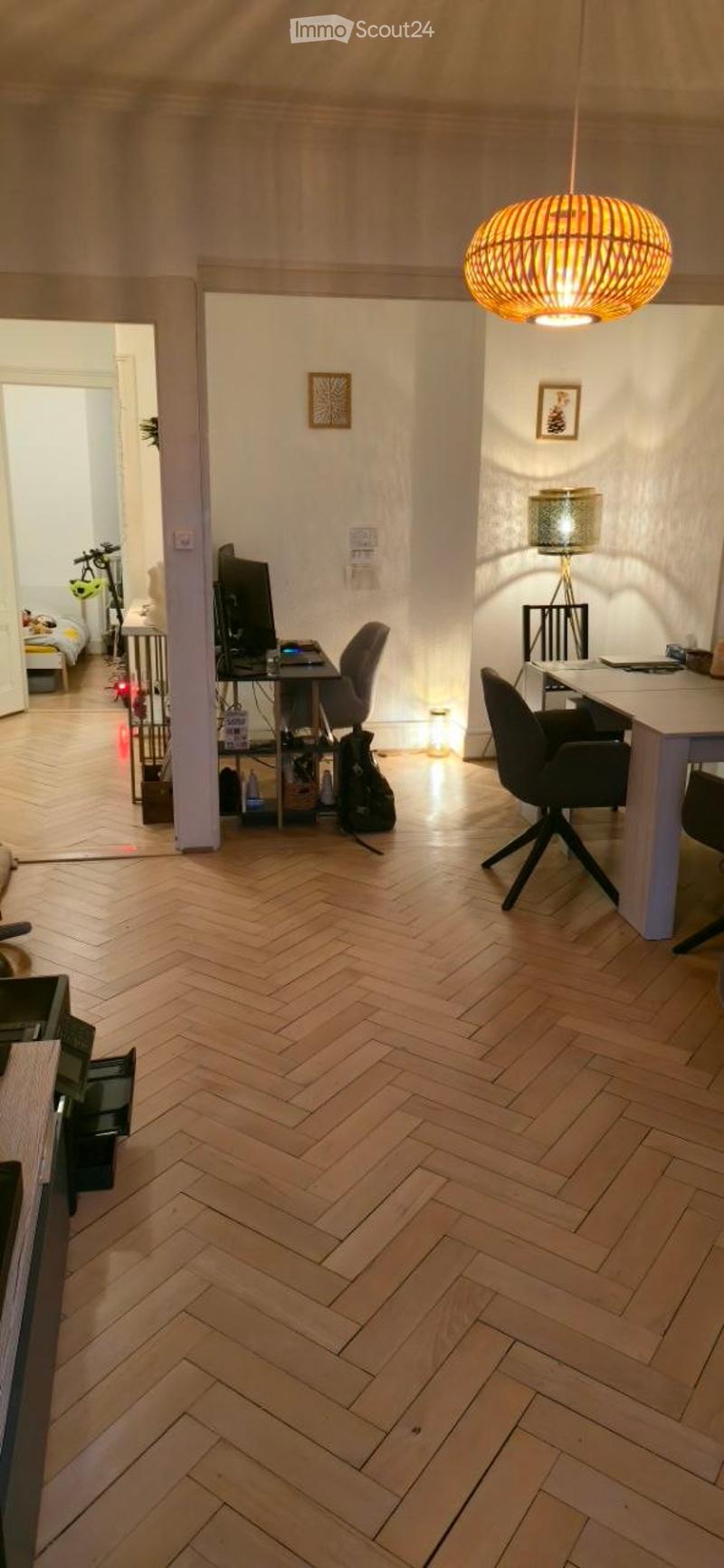 A modern, well-lit room with herringbone wooden flooring, a large desk with a computer screen, a comfortable chair, a round table with a lamp, and a hanging pendant light. The room is connected to another space with a bed visible in the background.