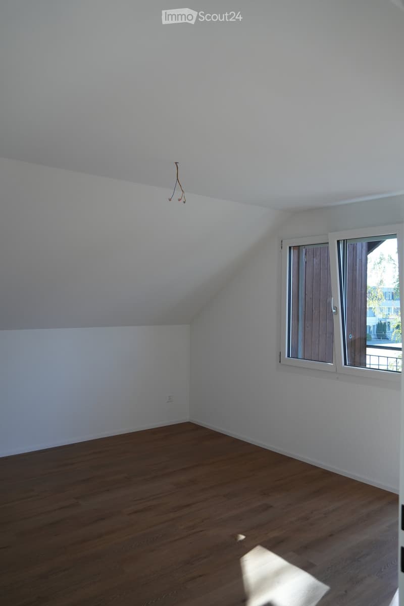 Empty attic room with wooden flooring, white walls, and a single window with blinds. The ceiling has a sloped design typical of attic spaces.
