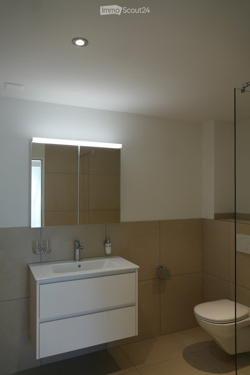Modern bathroom with a white sink, mirror, and toilet. The sink has a sleek design with a built-in cabinet below. The walls are tiled in neutral tones, and the floor is tiled in a warm, earthy color.