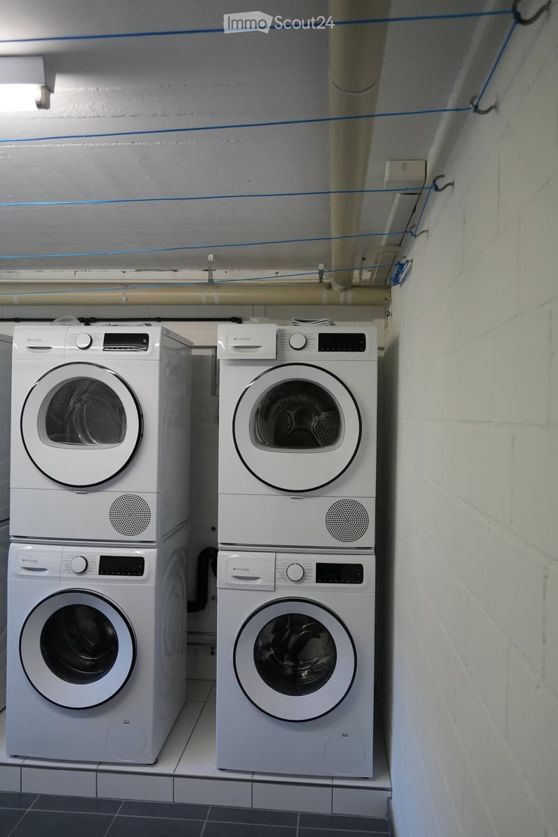 Two stacked washing machines and dryers in a utility room with exposed pipes and a tiled floor.