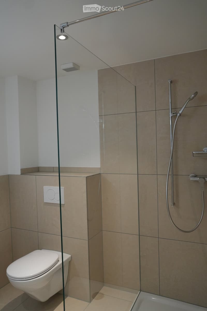 Modern bathroom with a toilet, glass-enclosed shower area, and a handheld showerhead. Beige tiles and a sleek design.
