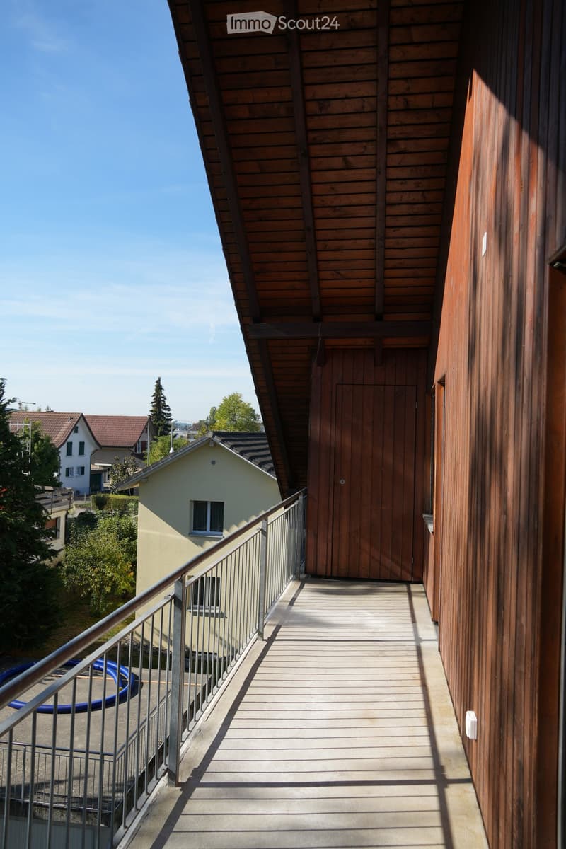 Wooden hallway with metal railing, leading to a wooden door, with a view of neighboring houses and greenery.