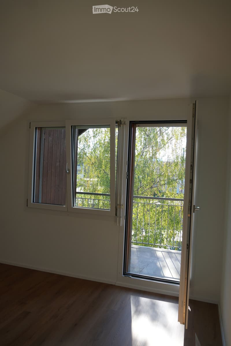 Empty room with wooden flooring, large sliding glass doors leading to a balcony with metal railing and greenery visible outside.