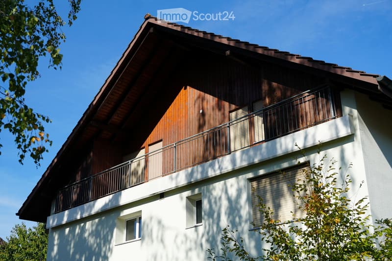 Two-story house with a wooden facade on the upper level and white walls on the lower level, featuring a balcony with metal railings and multiple windows.