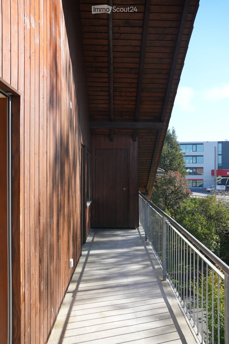 Wooden-walled corridor with a metal railing leading to a door, featuring a slanted wooden ceiling and natural lighting.