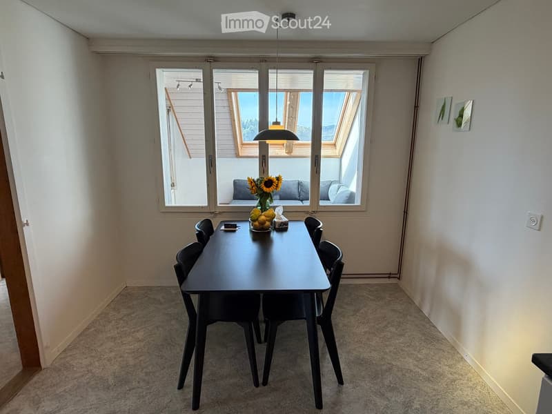 Dining room with a black dining table, six black chairs, a vase with sunflowers, and a fruit bowl on the table. Large windows with blinds and a view of the balcony area.