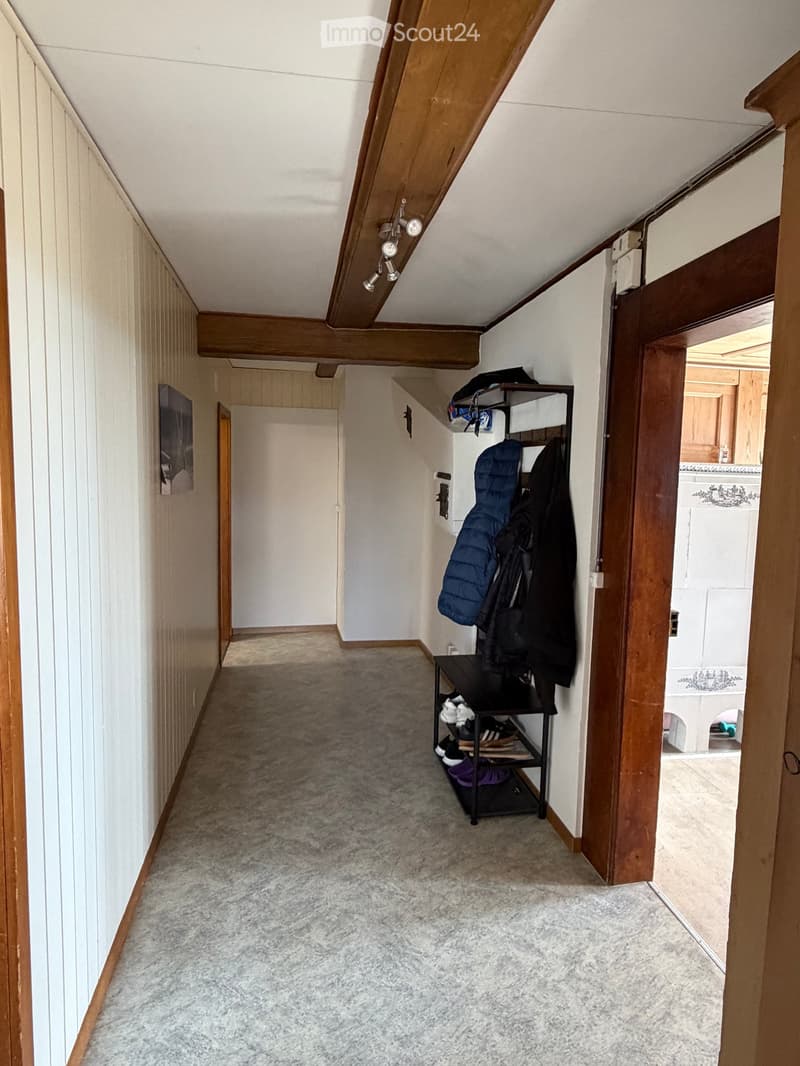 A narrow hallway with wooden beams on the ceiling, carpeted floor, and a coat rack with jackets and shoes. A small shelf holds additional items, and a doorway leads to another room.