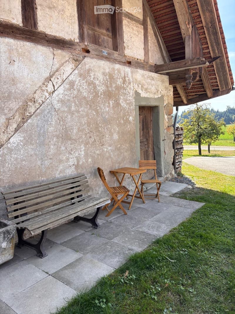 Outdoor seating area with a wooden bench, small table, and two folding chairs placed against a stone building with a wooden roof extension.