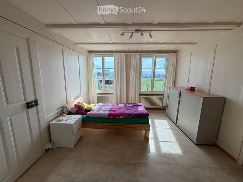 Bright bedroom with large windows offering a scenic view, featuring a bed with colorful bedding, a nightstand with a lamp, and a wardrobe.