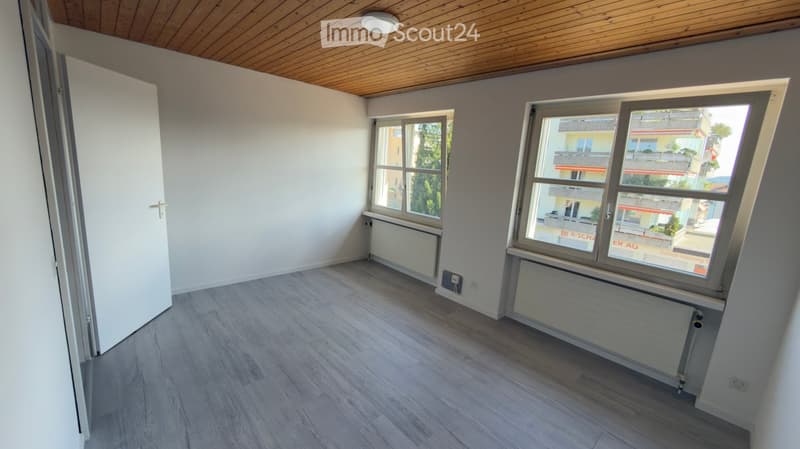 Empty room with light wooden flooring, white walls, and large windows offering a view of the street. The room features a wooden ceiling and radiators under the windows.