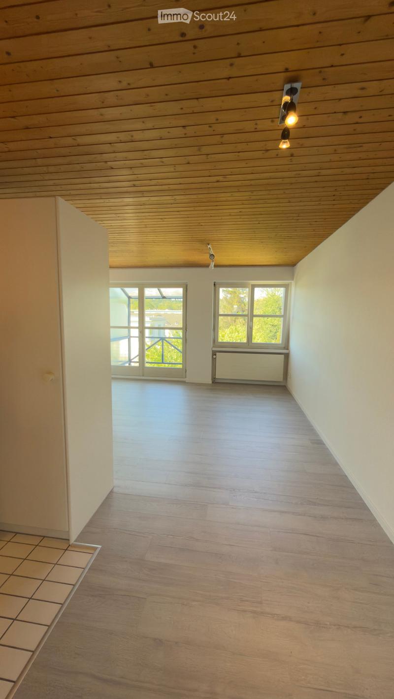 Empty room with wooden flooring, white walls, and a wooden ceiling. Features include a large window, a sliding glass door leading to a balcony, and a radiator.