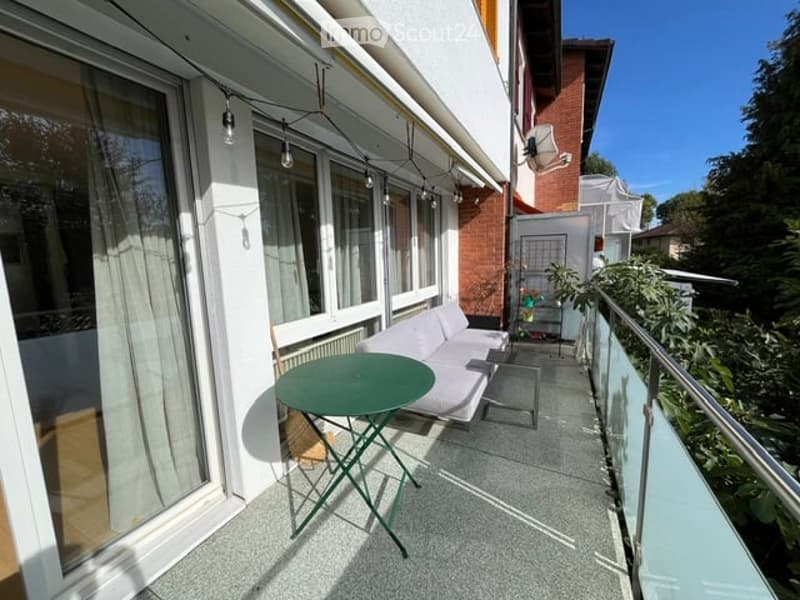 Balcony with a white sofa, green table, and glass railing, overlooking a garden area.