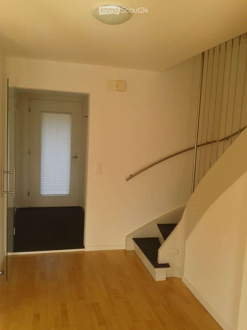 Empty hallway with wooden flooring, white walls, a staircase with a metal railing, and a glass door leading to another room.