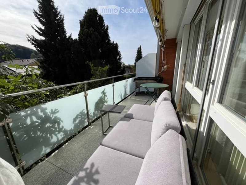 Balcony with a modern glass railing, a grey sofa, a small table, and a view of green trees and a house in the background.