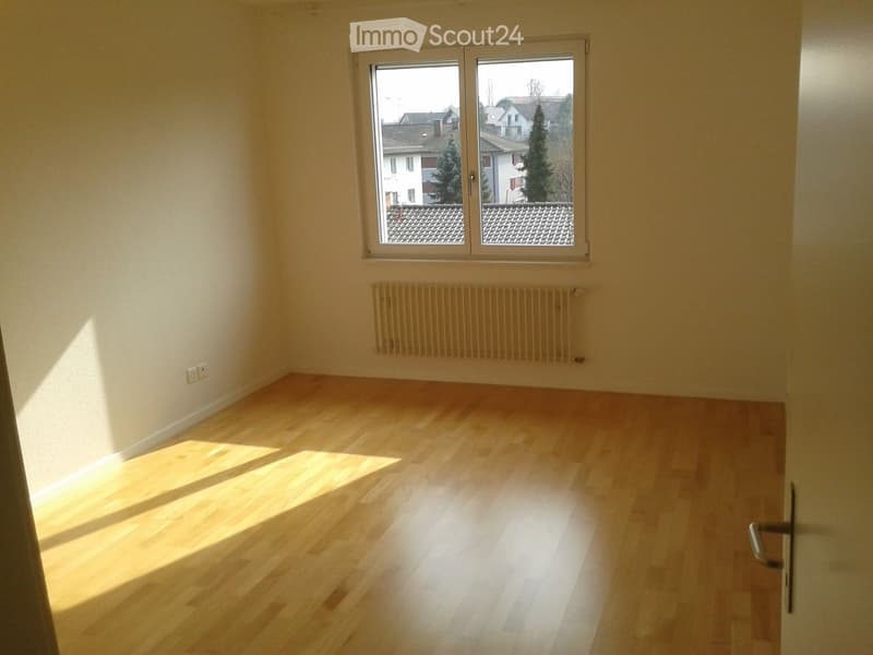 Empty room with wooden flooring, white walls, a window with a view of rooftops, and a radiator below the window.