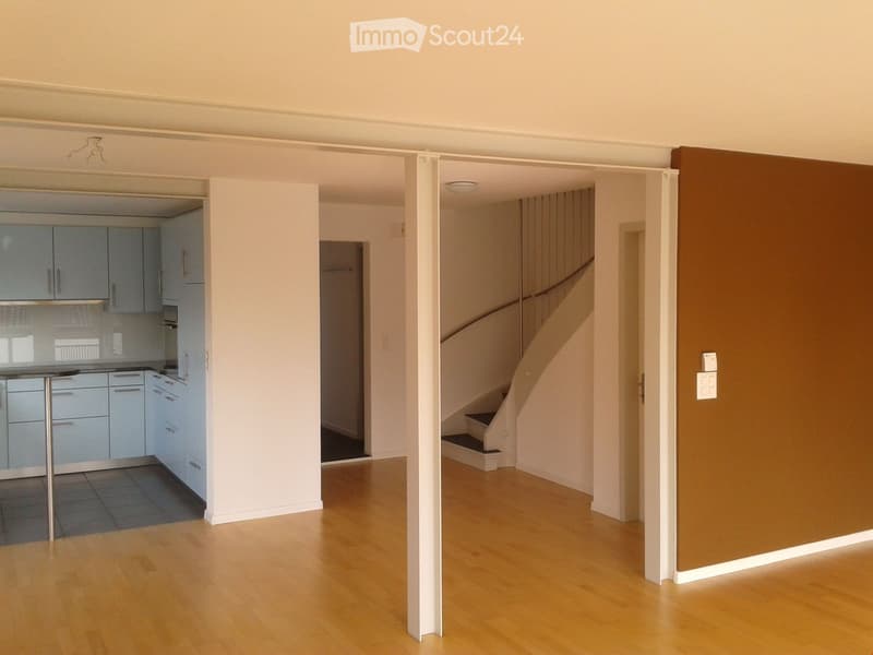 Bright, open hallway with wooden flooring, leading to a modern kitchen and staircase. The kitchen features light blue cabinets and stainless steel appliances.