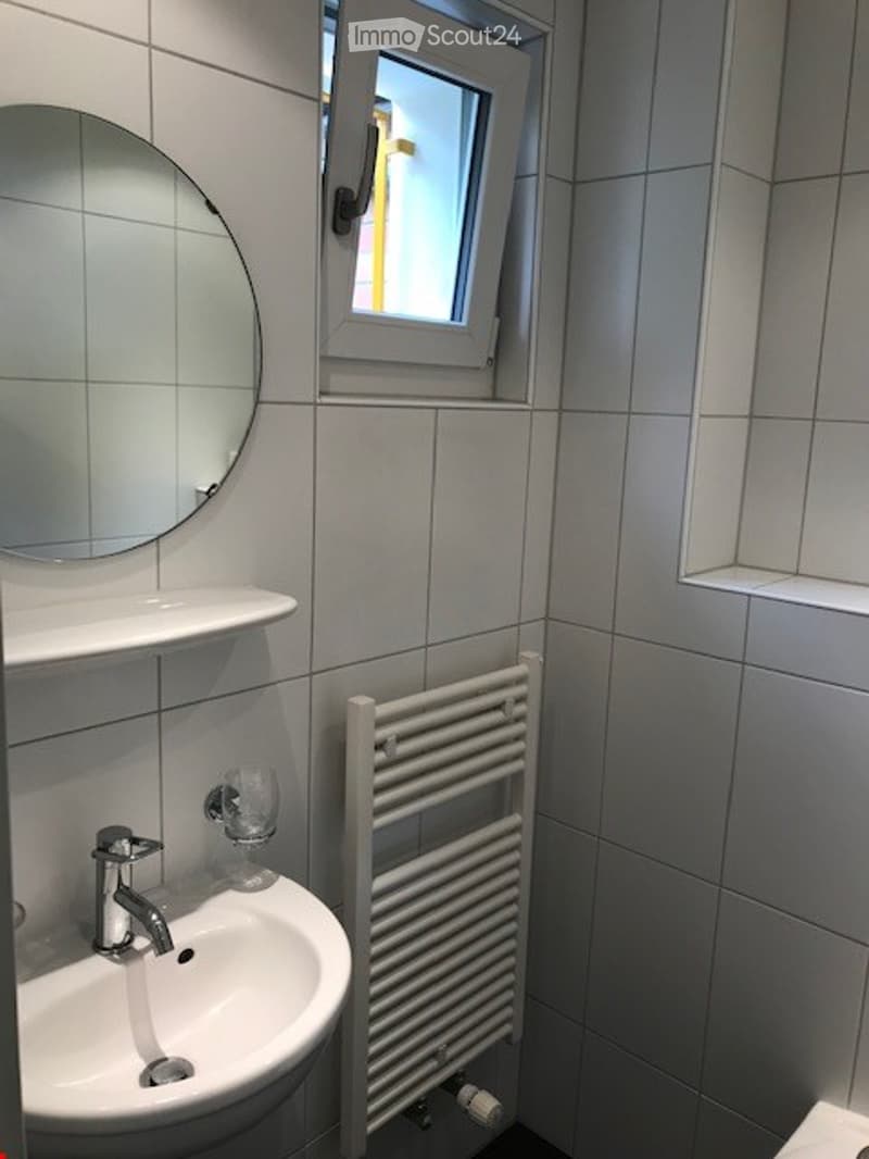 Modern bathroom with white tiled walls, a white sink with a chrome faucet, a round mirror, a white radiator, and a small window above the sink.