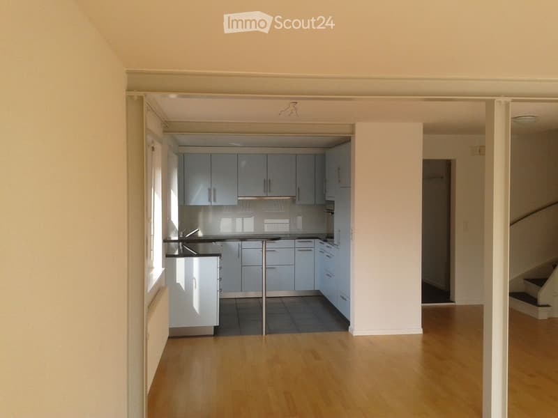Empty room with light wooden flooring, white walls, and a modern kitchen area visible in the background. No furniture or appliances are present.