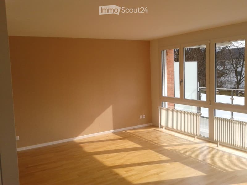 Empty room with light wooden flooring and large windows allowing natural light to fill the space. Beige walls and white radiators are visible.