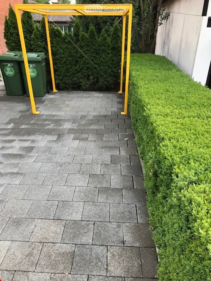 A paved outdoor area with neatly trimmed hedges, a yellow metal frame structure, and green waste bins.