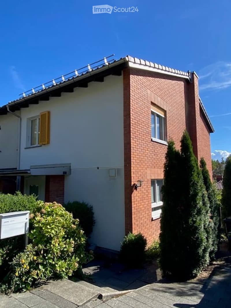 Two-story house with red brick facade, white walls, and yellow shutters. Features a balcony on the upper level and is surrounded by greenery.