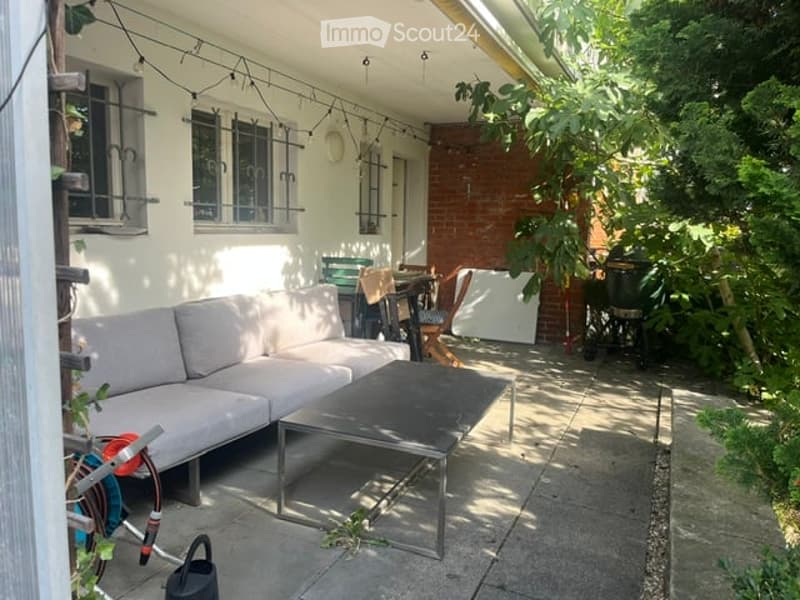 Outdoor patio area with a grey sectional sofa, black coffee table, and several chairs around a small table. The space is surrounded by greenery and has string lights overhead.
