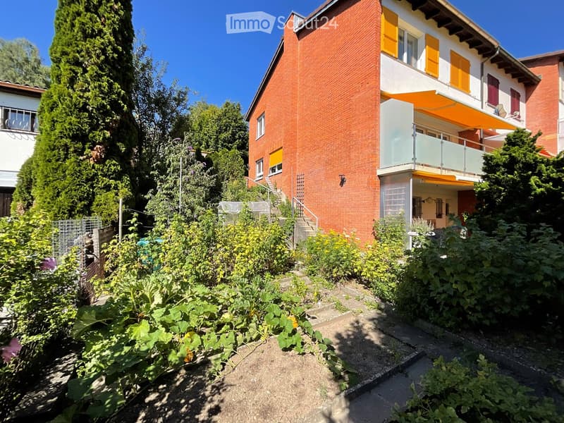 Two-story red brick house with yellow shutters, a balcony, and a well-maintained garden with various plants and a staircase leading to the entrance.