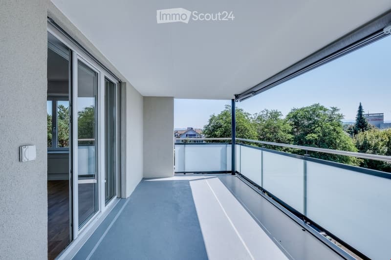 Modern balcony with white flooring, glass sliding doors, and a metal railing overlooking a green landscape.