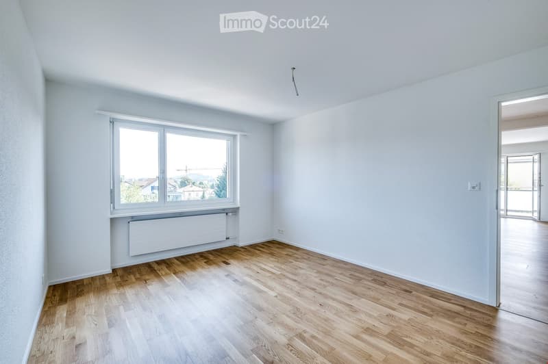 Empty room with light wooden flooring, white walls, large window with view, radiator, and open doorway leading to another space.