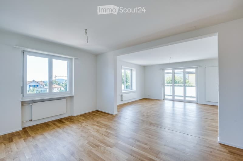 Empty room with light wooden flooring, white walls, and large windows offering a view of the outside. Radiators are visible under the windows.