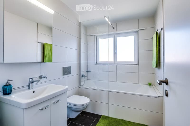 Modern bathroom with white tiles, a sink with a chrome faucet, a toilet, a bathtub with a glass shower enclosure, a large mirror, and a green towel hanging on the wall.