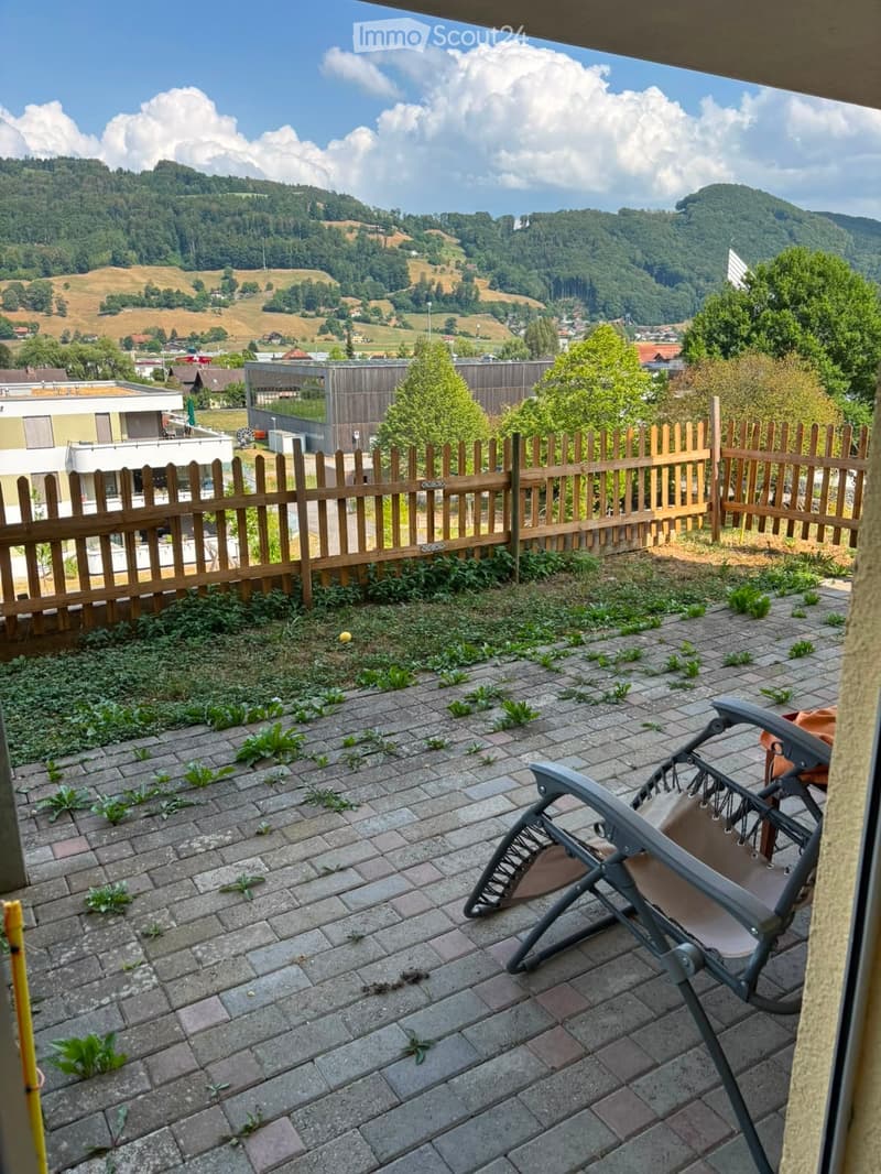 Balcony with brick pavement, wooden fence, and a single lounge chair, overlooking a scenic valley with hills and scattered buildings.