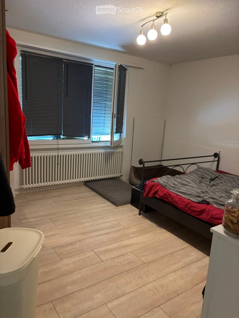 Bedroom with tiled floor, large window with blinds, radiator, bed with red and grey bedding, nightstand with jar, and a white trash bin.