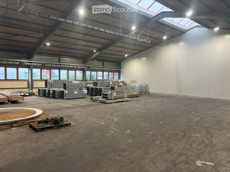 Large, empty industrial-style warehouse space with high ceilings, exposed beams, and large windows. The floor is concrete, and there are some stacked materials and pallets in the background.