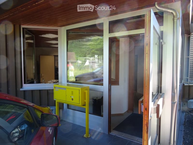 Entrance area of a building with a covered porch, large windows, a yellow mailbox, and a red car parked nearby.