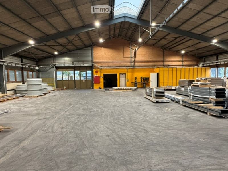 Large, empty industrial-style warehouse with a high ceiling, concrete floor, and scattered storage items including metal drums, stacked materials, and a few wooden pallets. The space has large windows and a skylight, with minimal furnishings.