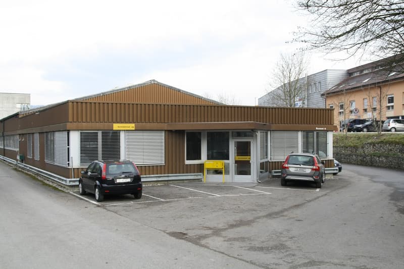 A modern, single-story building with a brown wooden facade and white trim, featuring large windows and a central entrance. Two cars are parked in front of the building.