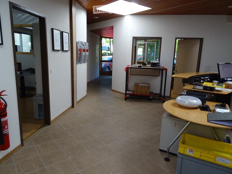 Office space with tiled floor, wooden ceiling, and several desks with computers, printers, and office supplies. Includes a fire extinguisher, framed pictures on the wall, and a cart with boxes.