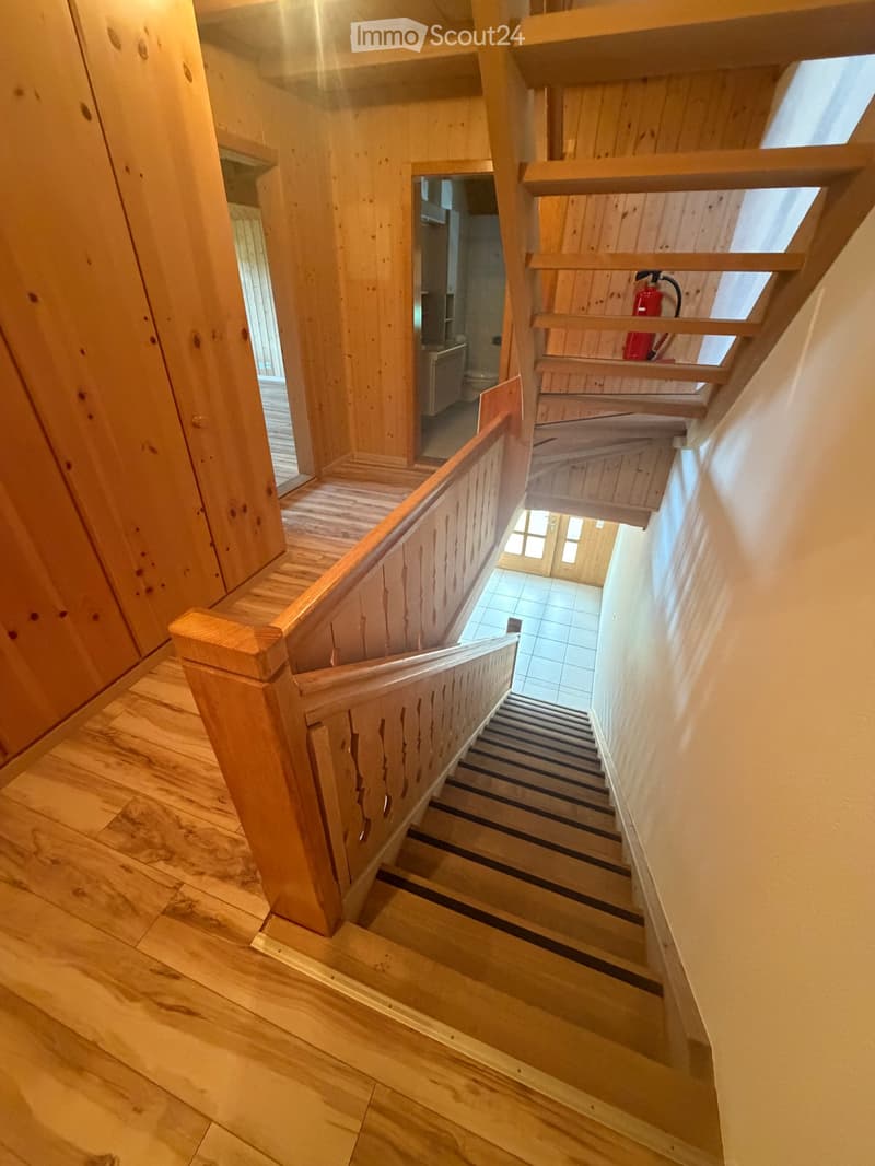 Wooden staircase with a carved railing, leading to a hallway with wooden walls and a tiled floor. A red fire extinguisher is visible on a shelf in the background.