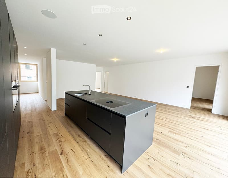 Modern kitchen with a central island, dark cabinets, and a stainless steel sink. The floor is made of light wooden planks, and the walls are white.
