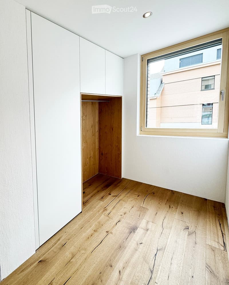 Empty room with light wooden flooring, white walls, built-in white cabinets, and a large window showing a neighboring building.