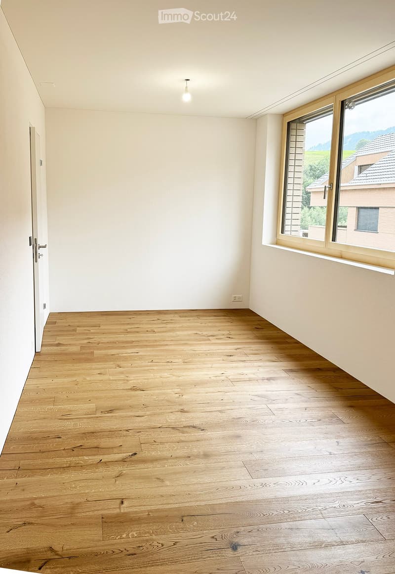 Empty room with light wooden flooring, white walls, and a large window with a view of the outdoors.