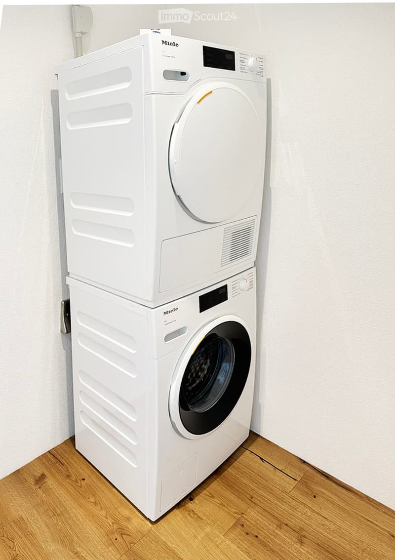 A white washing machine and dryer stacked vertically in a room with wooden flooring and white walls.