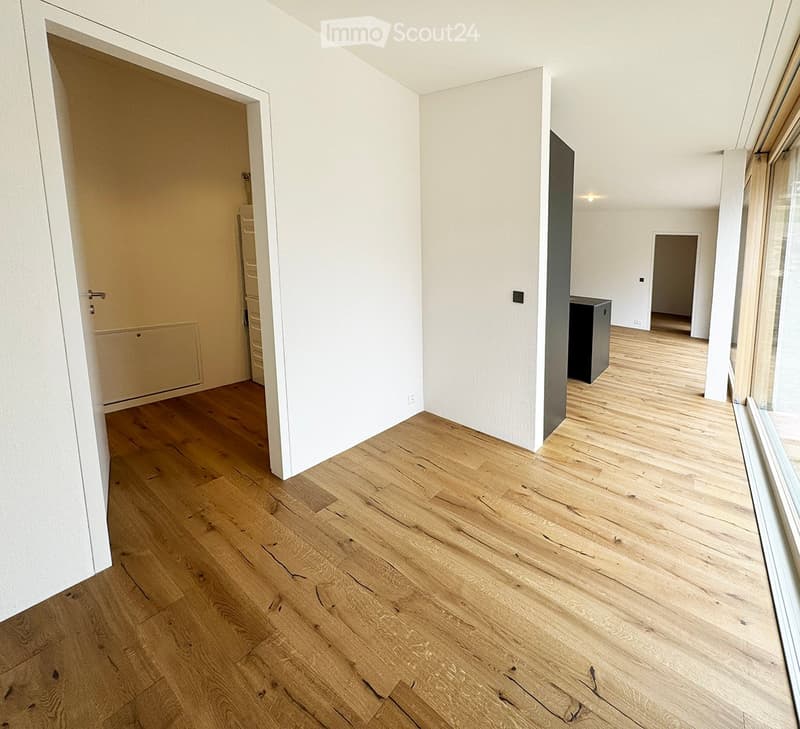 Empty hallway with light wooden flooring, white walls, and large windows allowing natural light. No furniture or personal items are present.