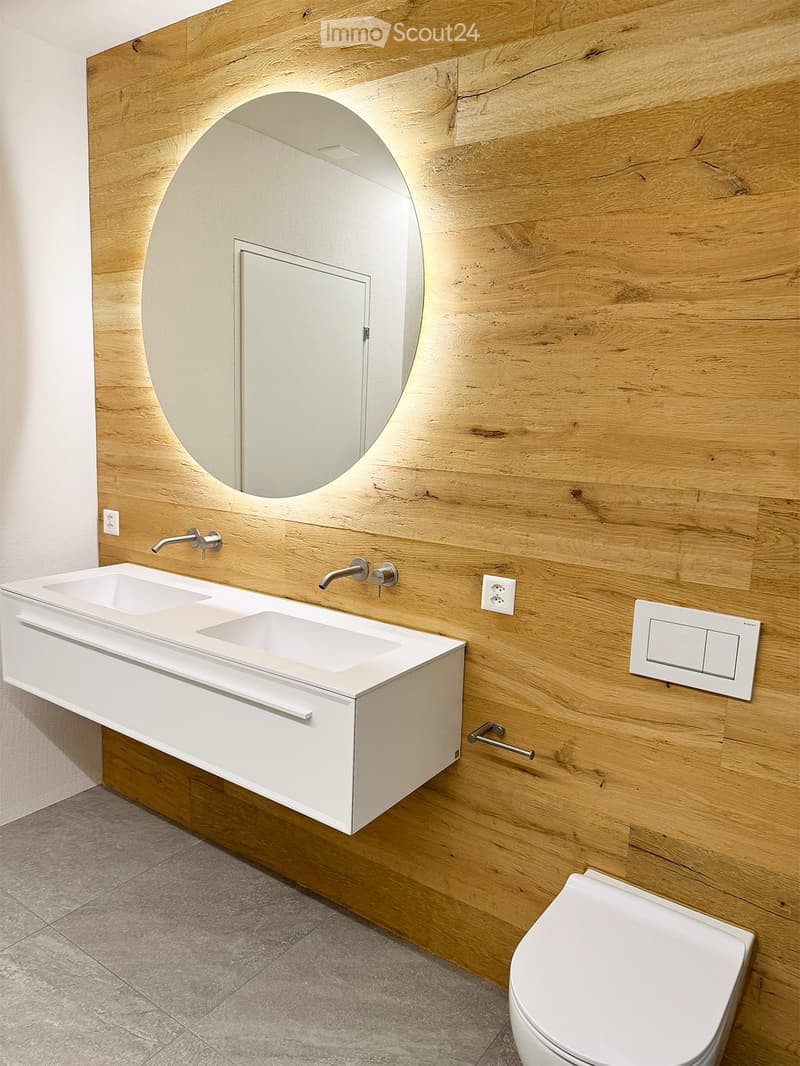 Modern bathroom with a white sink, dual faucets, a large round mirror, and a toilet, set against a wooden wall.