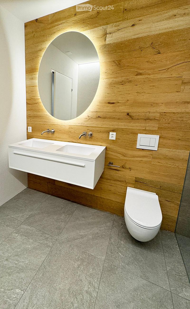 Modern bathroom with a white double sink, a toilet, and a large round mirror mounted on a wooden wall.